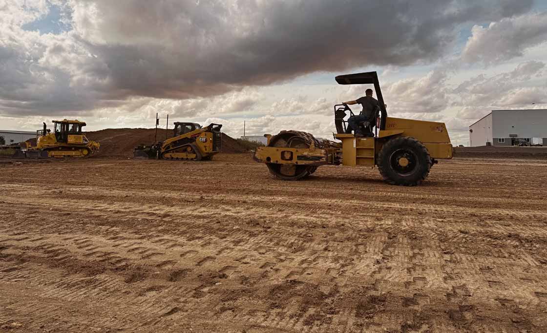 A man operating a yellow road roller on a large dirt construction site with two other yellow construction vehicles and a white building in the background under a cloudy sky.