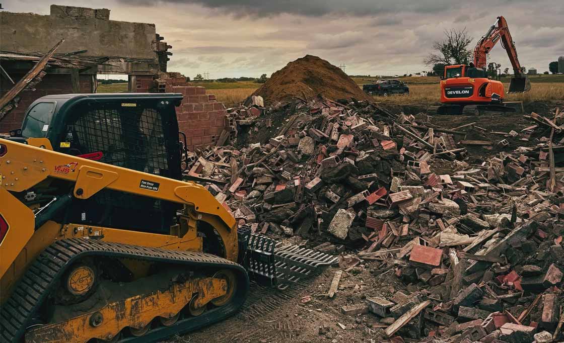 Construction site with two excavators and a large pile of rubble and dirt near a partially demolished brick wall.