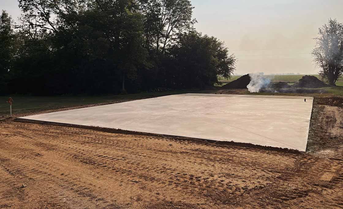Freshly poured concrete slab foundation on leveled dirt with trees and smoking dirt piles in the background.
