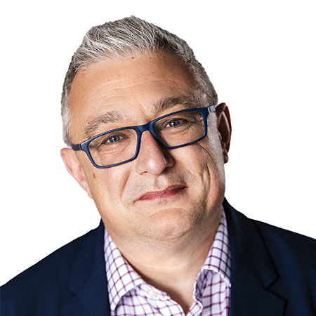 Smiling person with short white hair and wire-framed glasses, wearing a black suit jacket, white button-up, and a yellow-patterned tie