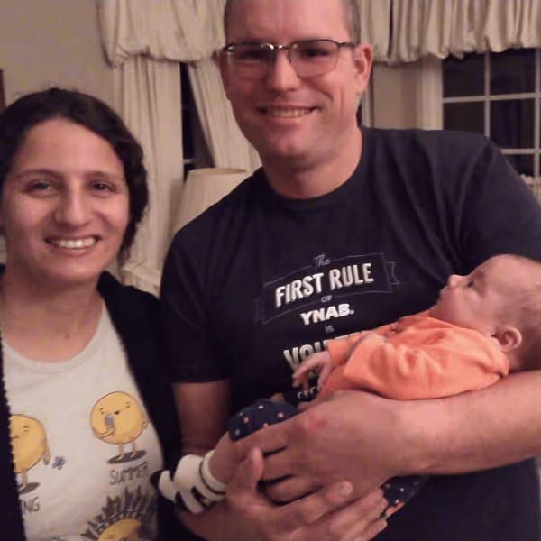 Smiling man holding a baby in orange clothes and woman standing beside him indoors.