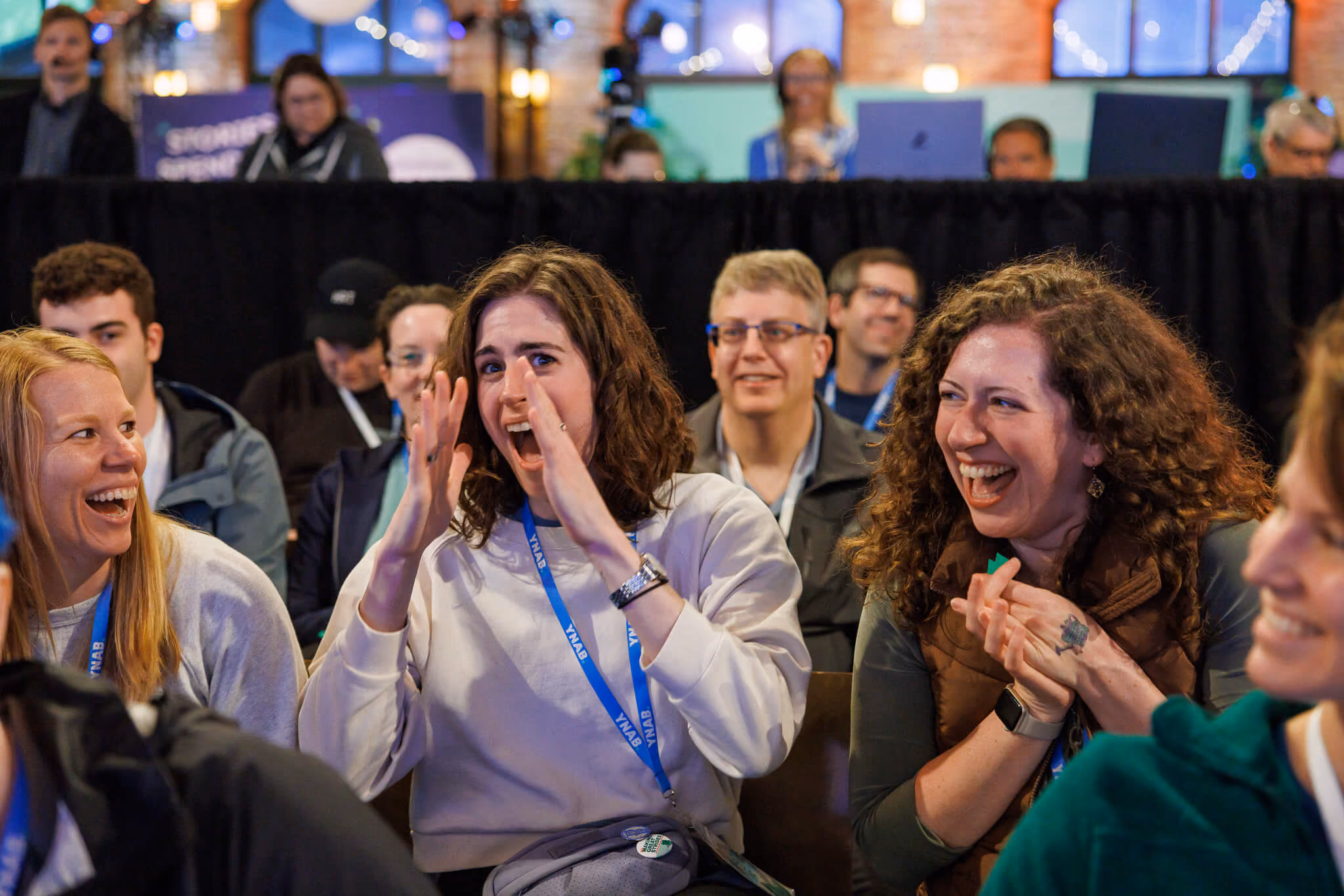 A group of people sitting indoors, smiling and laughing together, with some wearing blue lanyards at YNAB Fan Fest.