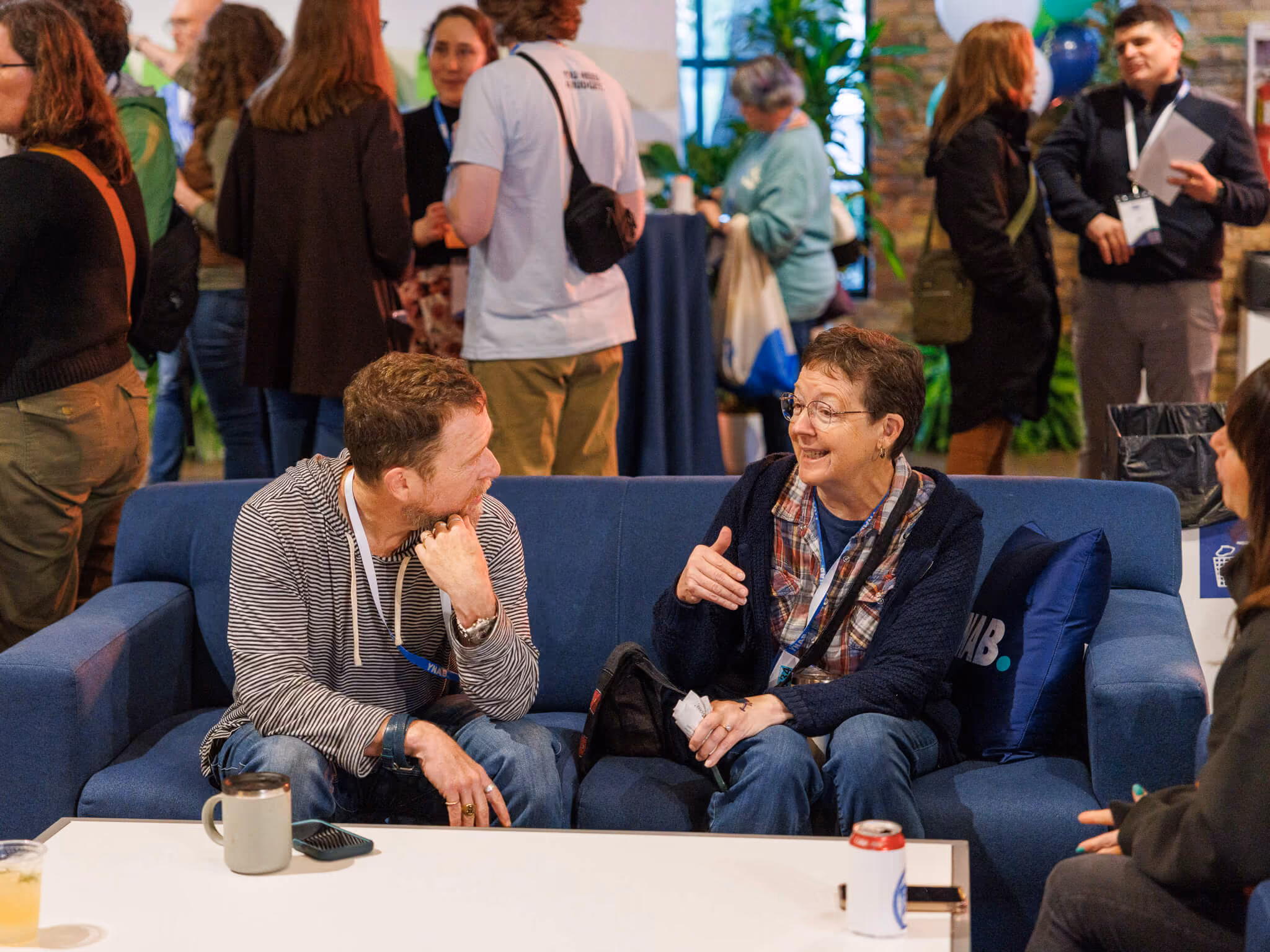 Two people sitting on a blue couch engaged in a friendly conversation in a busy indoor event space with others standing and mingling in the background.