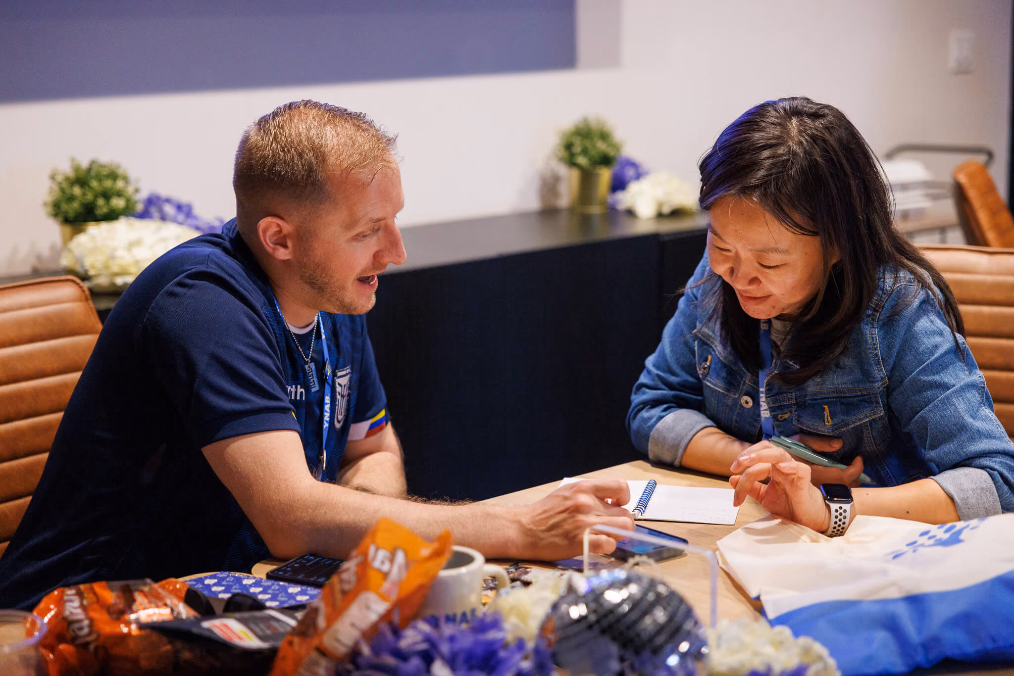 A man and a woman sitting at a table engaged in a conversation or discussion, with papers, a notebook, and snacks on the table in front of them.
