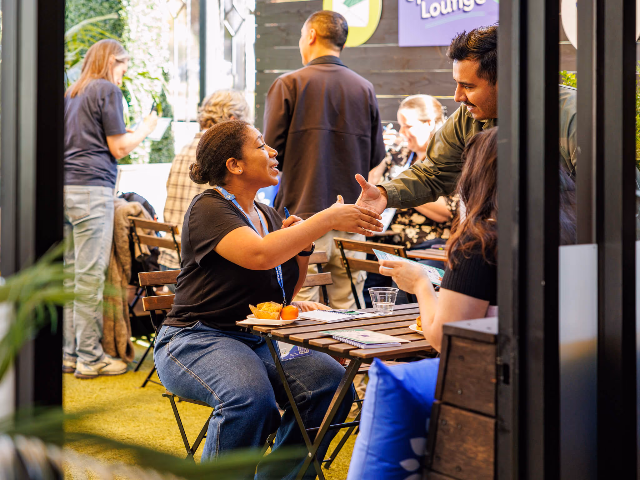 Two people sitting at an outdoor table warmly shaking hands while others socialize in the background at a casual gathering.