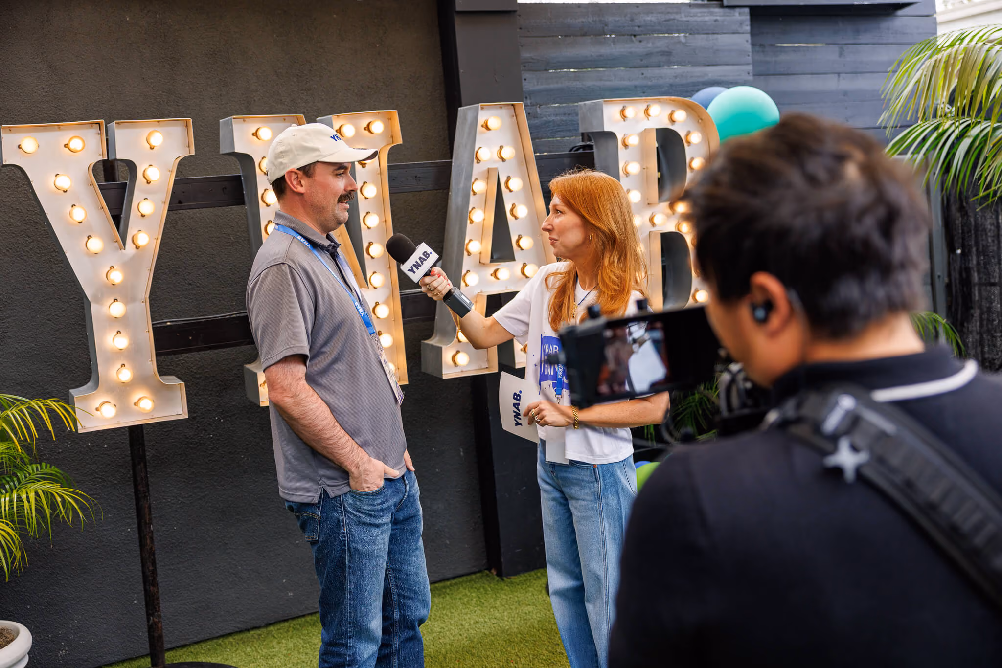 A woman with red hair holding a microphone labeled YNAB interviews a man wearing a gray polo shirt and a light gray cap, while a cameraman films them in front of large light-up letters spelling YNAB on a black wall with green plants nearby.