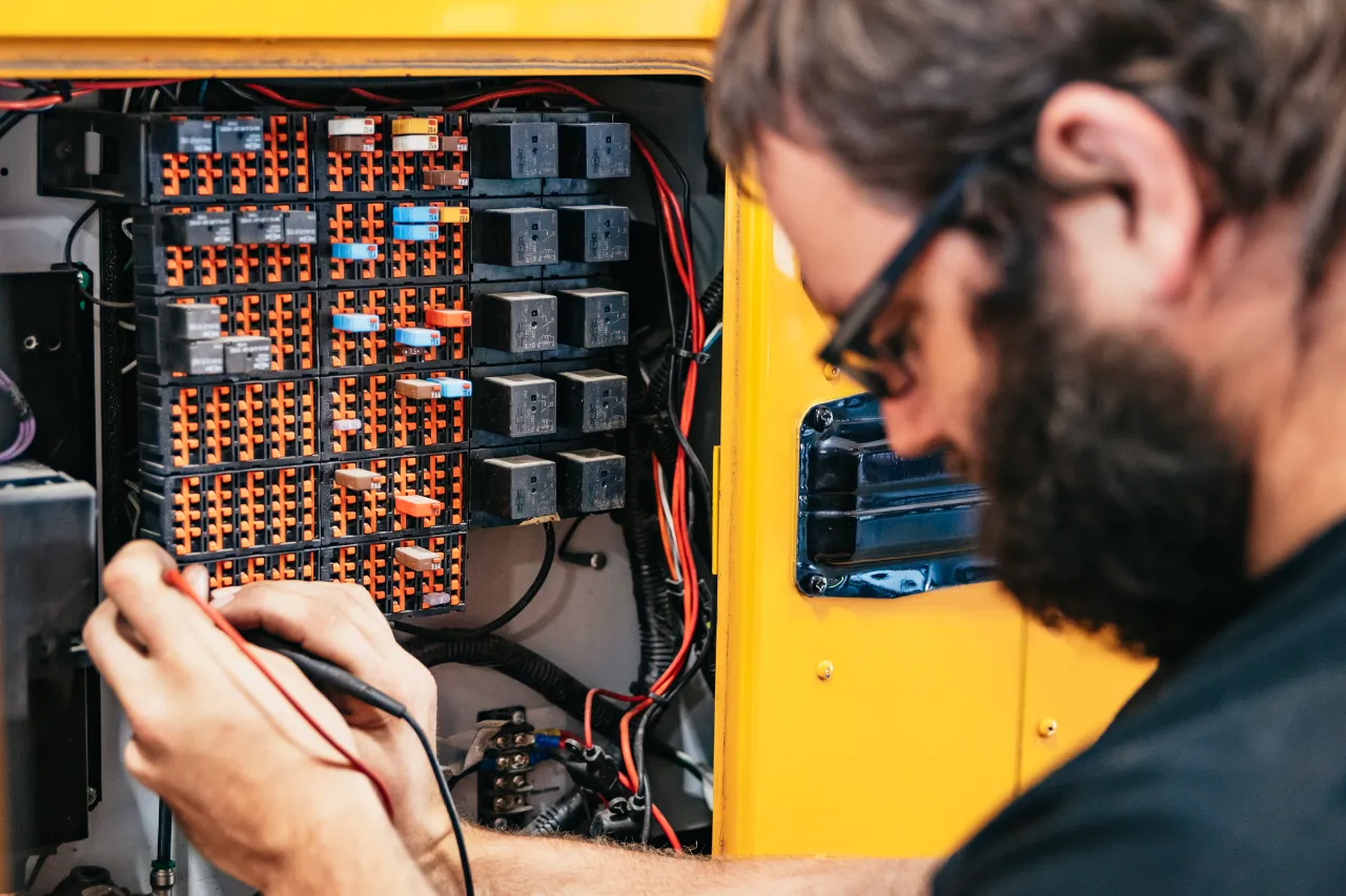 Diesel repair partner testing electrical circuits beside a panel of relays and fuses inside a yellow service vehicle.