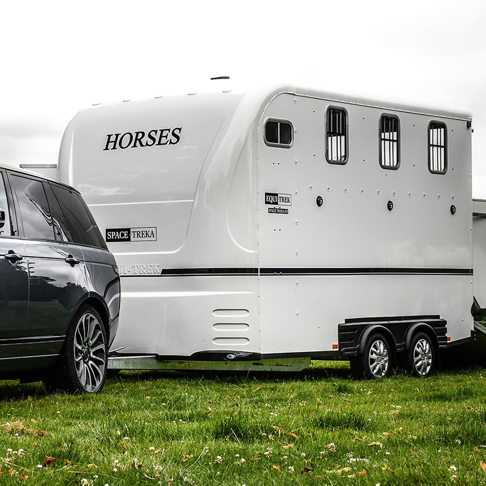 A large white horse trailer labeled "Equi-Trek Space-Treka" being towed by a dark grey SUV on a grassy field. The trailer features a curved aerodynamic front, barred windows for ventilation, and dual axles with chrome wheels.