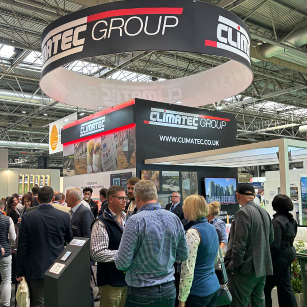 A bustling trade show exhibit for Climatec Group inside a large convention hall. A large, circular black and red banner with the company logo hangs from the ceiling over the booth. Several attendees are gathered in the foreground, engaged in conversation near a black display wall that features the company's website, "www.climatec.co.uk," and images of agricultural environmental control equipment.