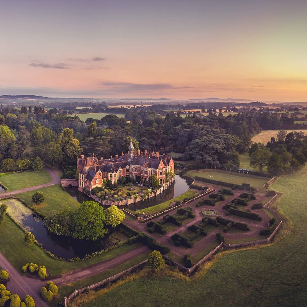 An aerial sunset view of Madresfield Court, a moated stately home in Worcestershire, England. The historic red-brick manor is surrounded by a calm moat, meticulously manicured hedge gardens, and sprawling green parkland. The warm evening light casts long shadows across the landscape, with rolling hills visible in the hazy distance under a soft orange and purple sky.