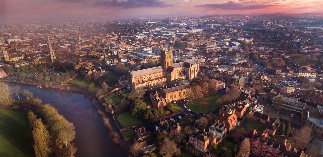 The magnificent Worcester Cathedral stands as the dramatic centerpiece. Its massive, sandy-brown stone structure features intricate Gothic architecture, a prominent central tower, and sprawling monastic buildings that border a patch of vibrant green lawn.