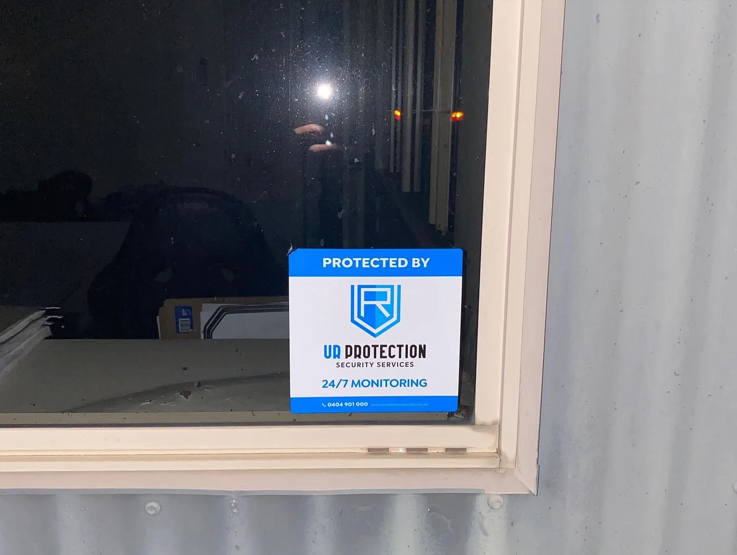 Window with a blue and white sticker that reads 'Protected by UR Protection Security Services 24/7 Monitoring' with contact details at the bottom.