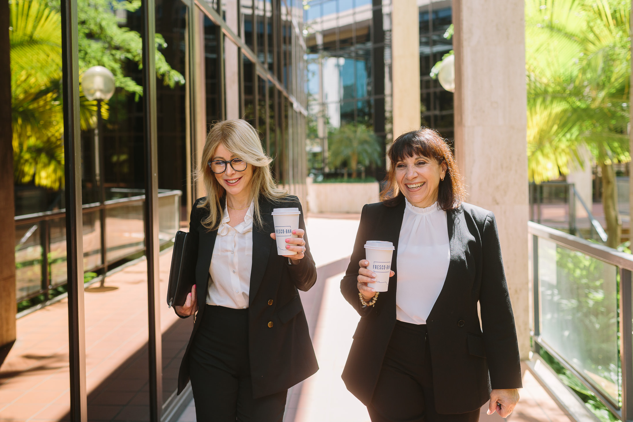 SA brand photography - Two female colleagues , walk together to a business meeting enjoying a coffee and smiling with each other. Palms photography, Adelaide commercial photographer