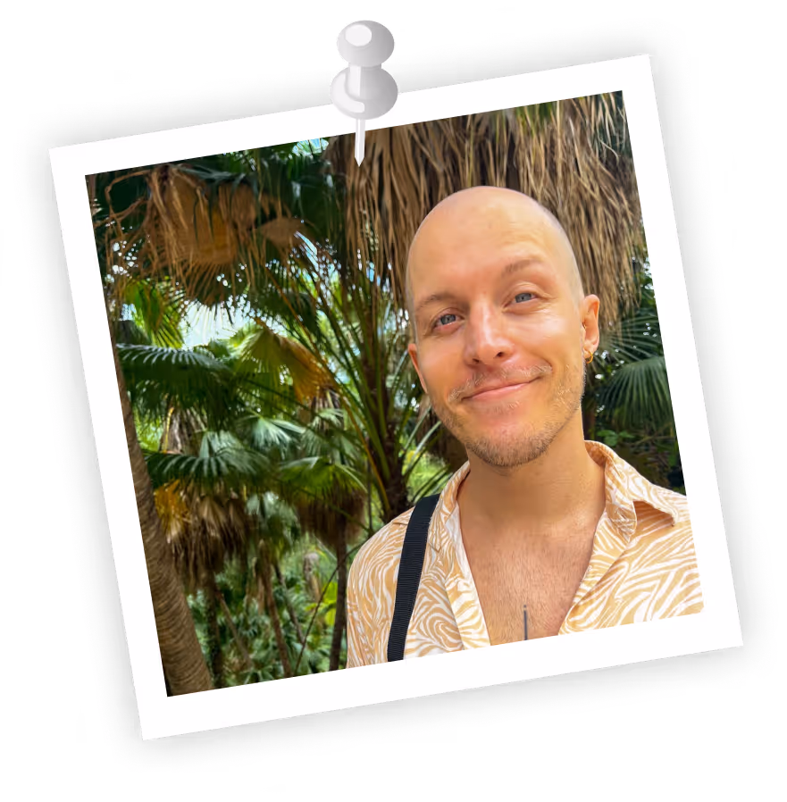 Smiling bald man wearing a patterned shirt standing in front of lush green palm trees.