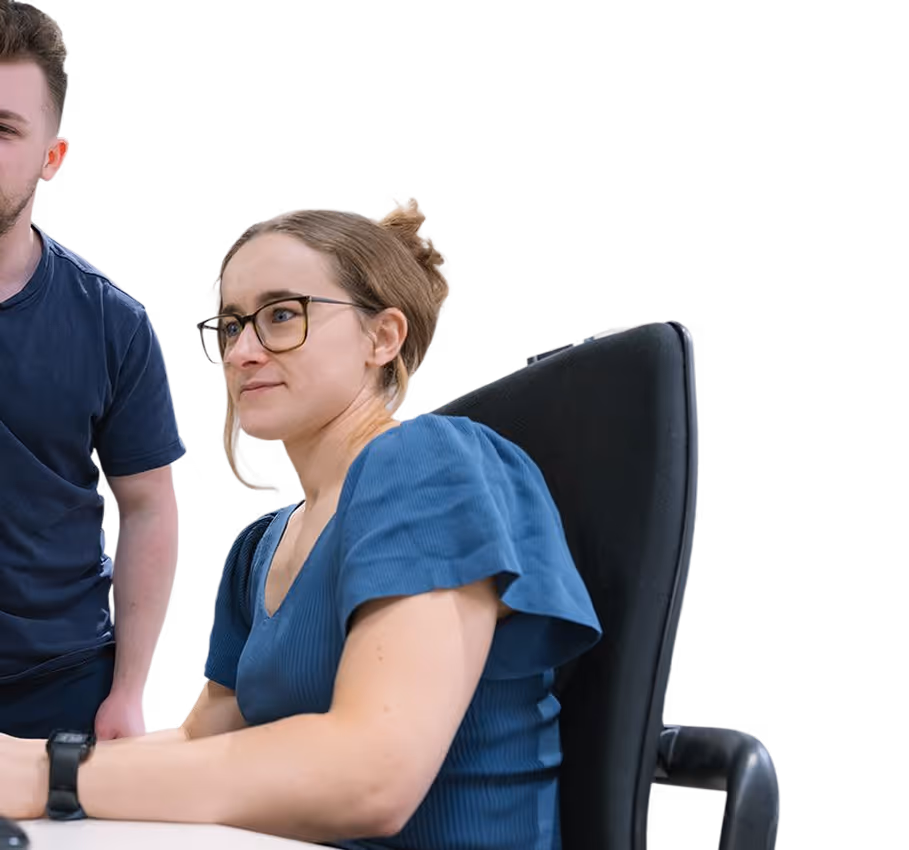 Woman with glasses in blue dress sitting on black chair at white desk, man in dark shirt stands beside her.