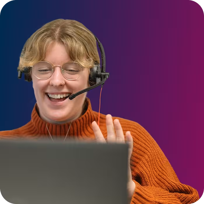 Smiling person with glasses and headset waving while looking at a laptop screen.