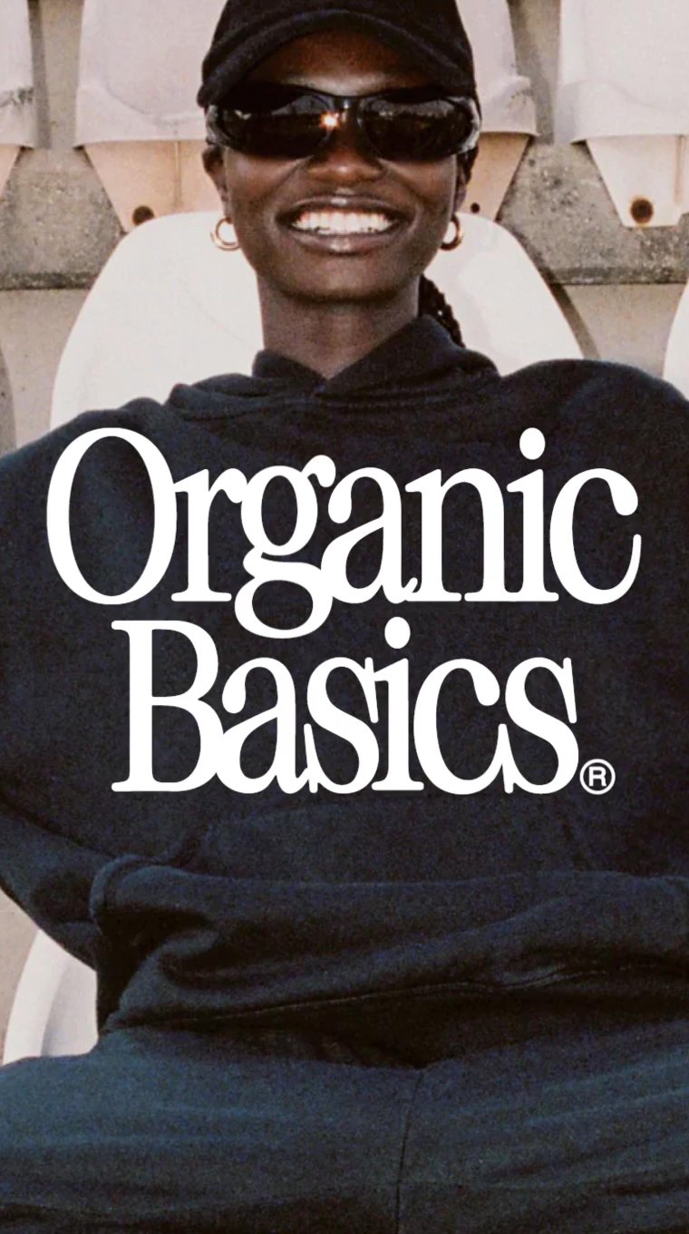 Smilende person med mørke solbriller og kasket iført sort hættetrøje med teksten Organic Basics.