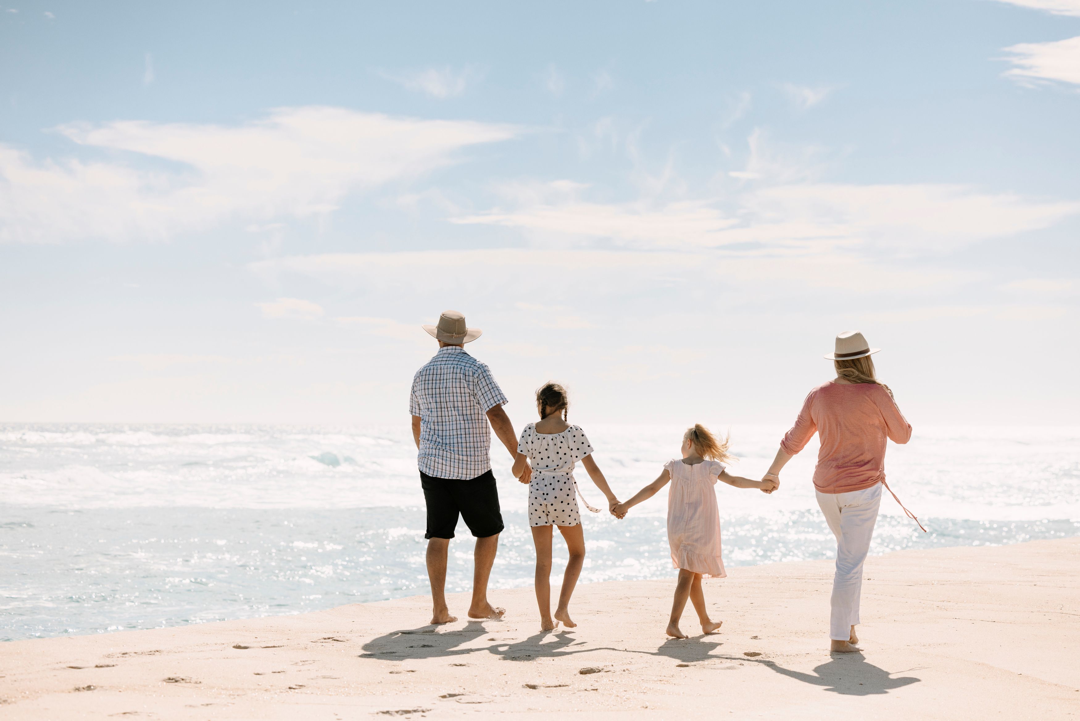 Family running into the ocean together, looking back at camera and smiling. Family includes grandparents, parents and a boy and girl. Weather is sunny with slight cloud and the water is clear and slightly wavey.