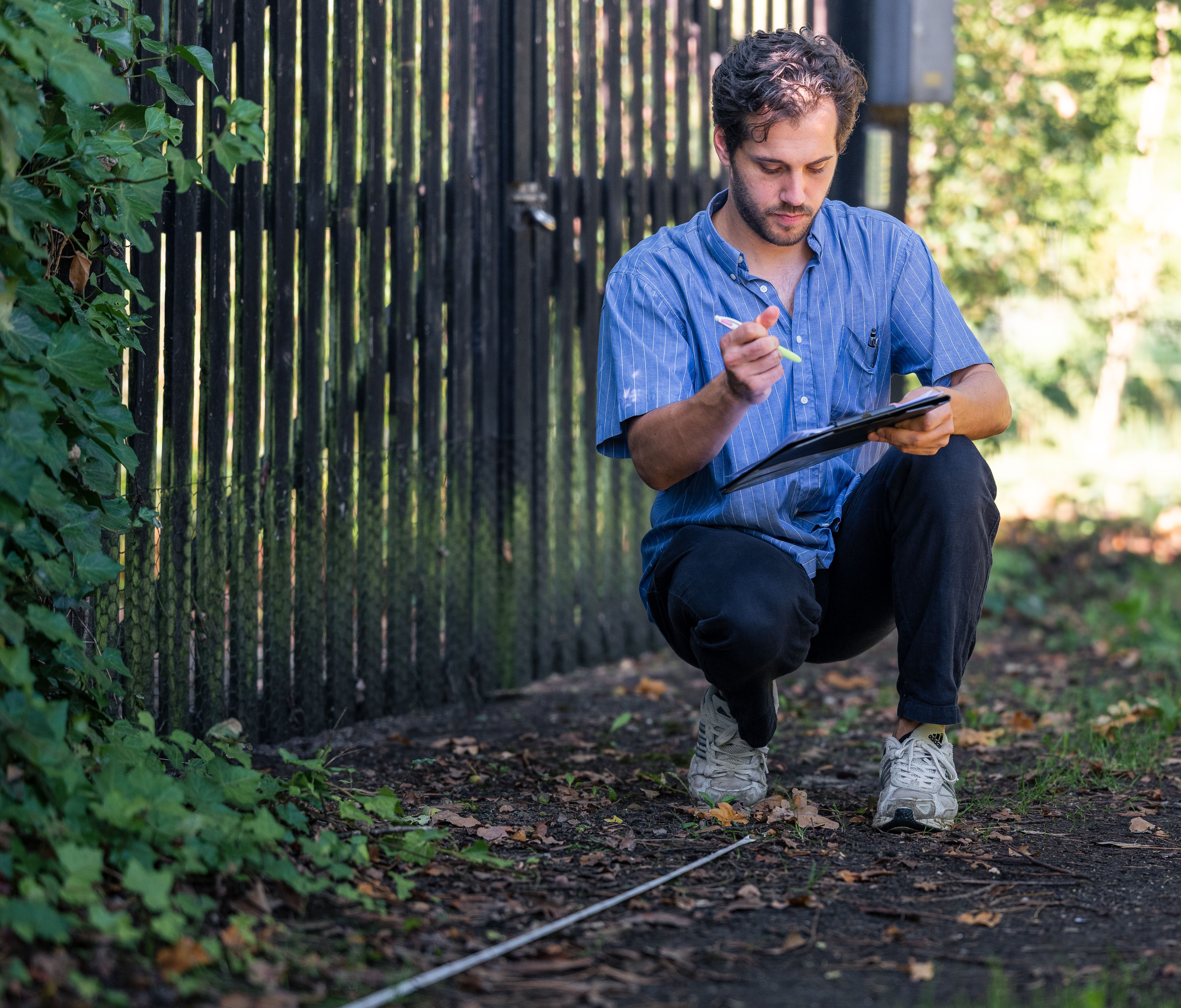 Man knielt buiten bij een metalen hek en meet met een meetlint terwijl hij notities maakt op een klembord.