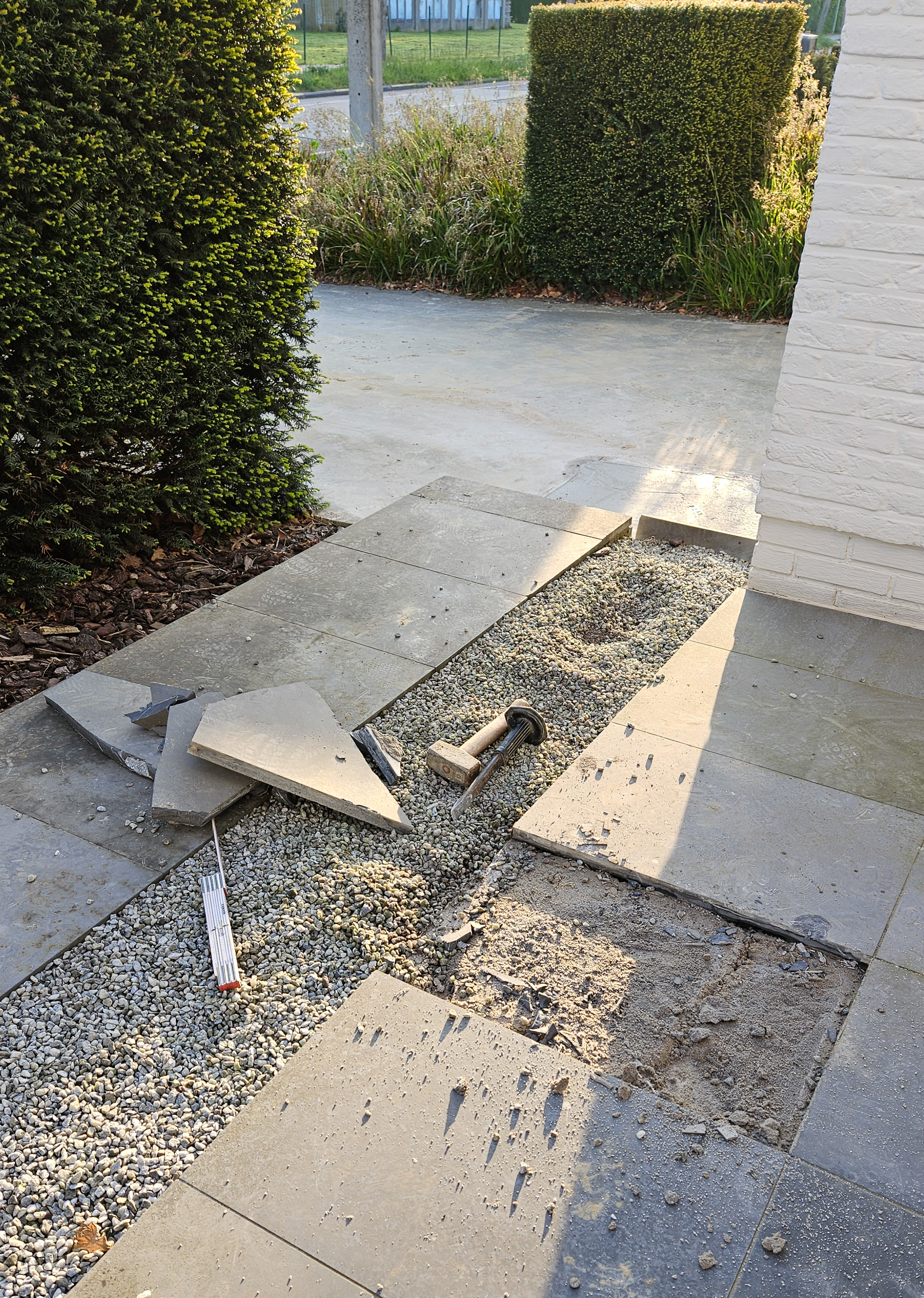 Buitenbetegeling deels verwijderd met gereedschap en grind zichtbaar bij een huis met groene heggen.