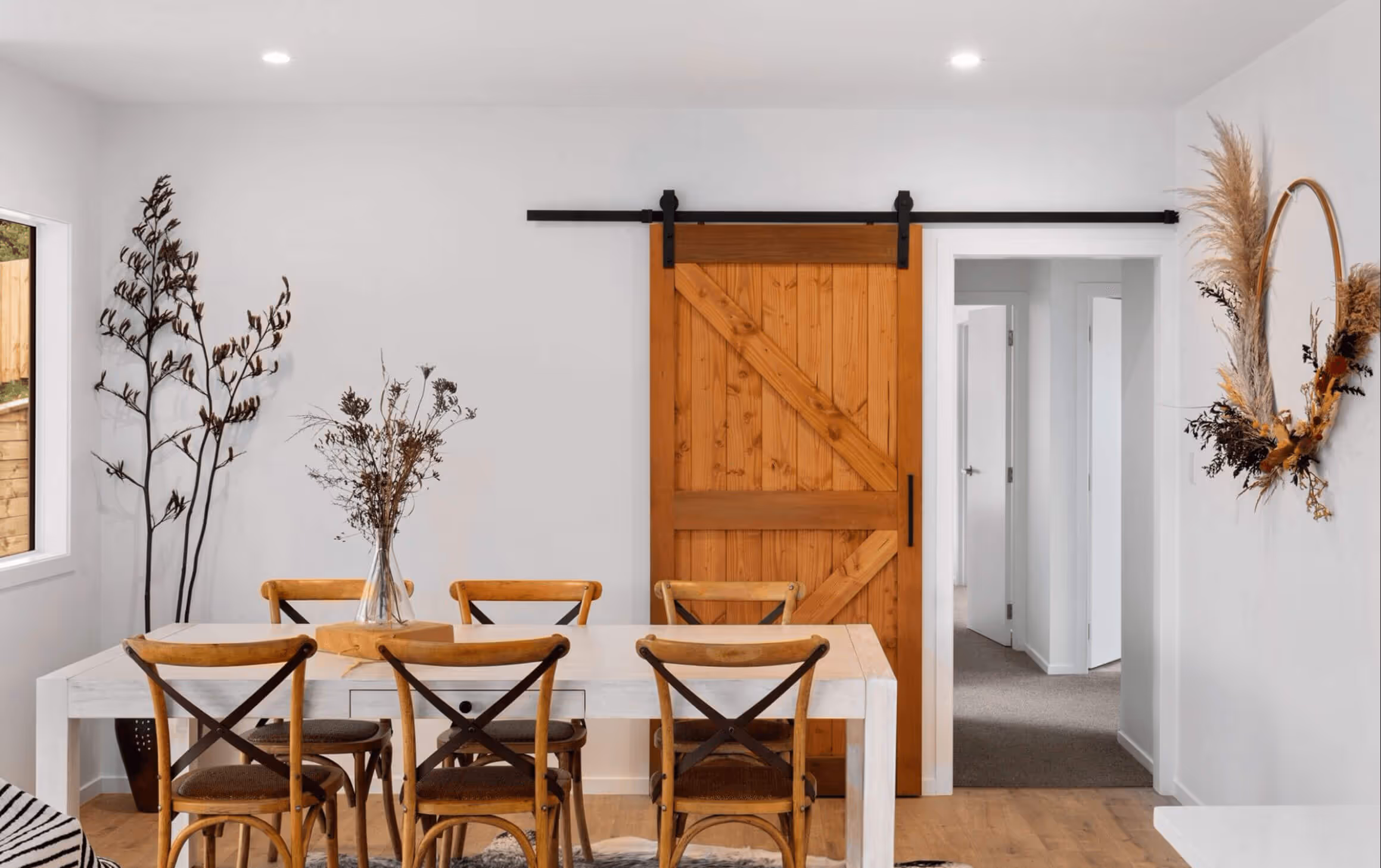 Dining area with a white table, six wooden chairs, a vase with dried flowers, and a wooden sliding barn door.