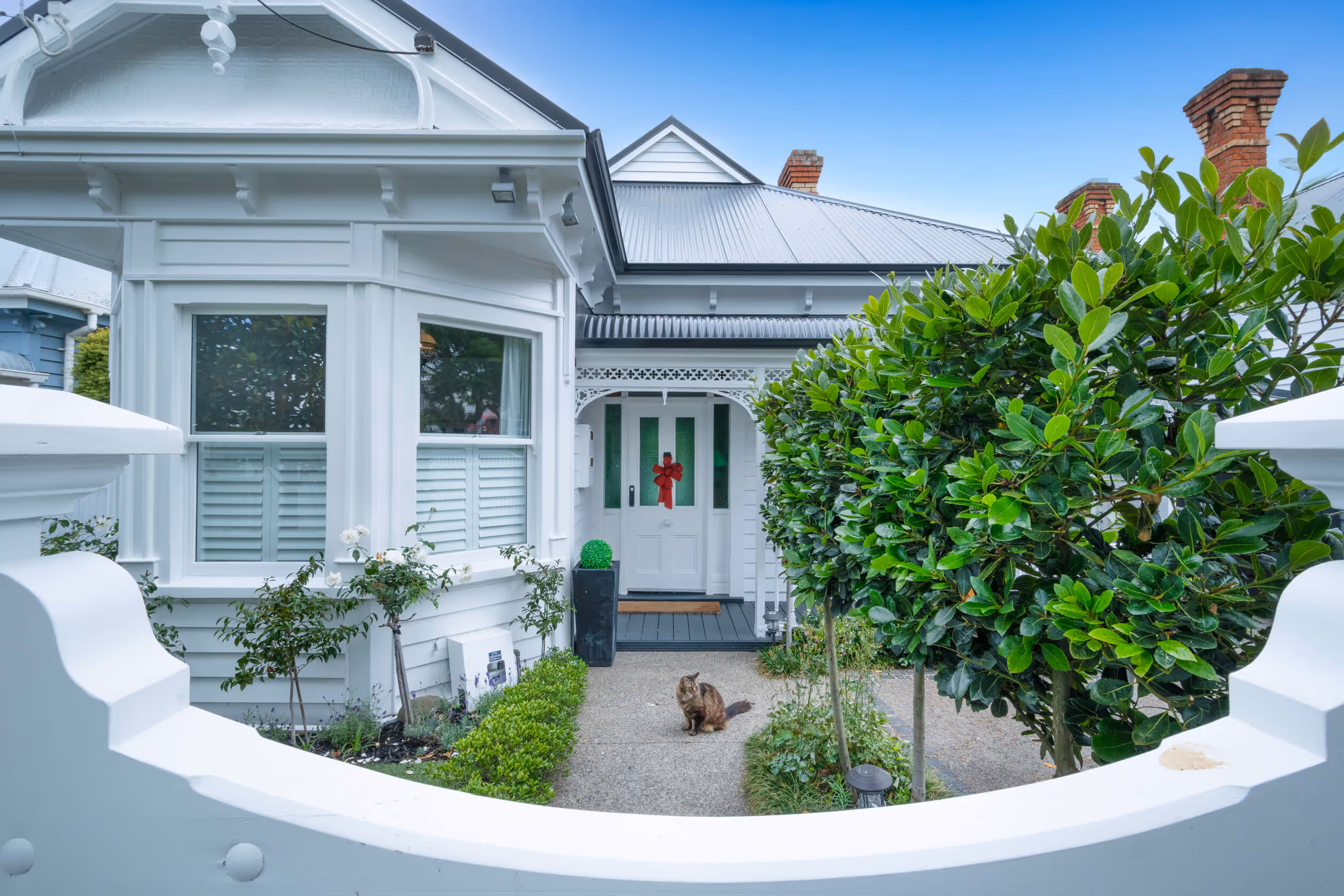 Villa house entrance with bay windows, a red bow on the front door, green bushes, and a cat sitting on the pathway.
