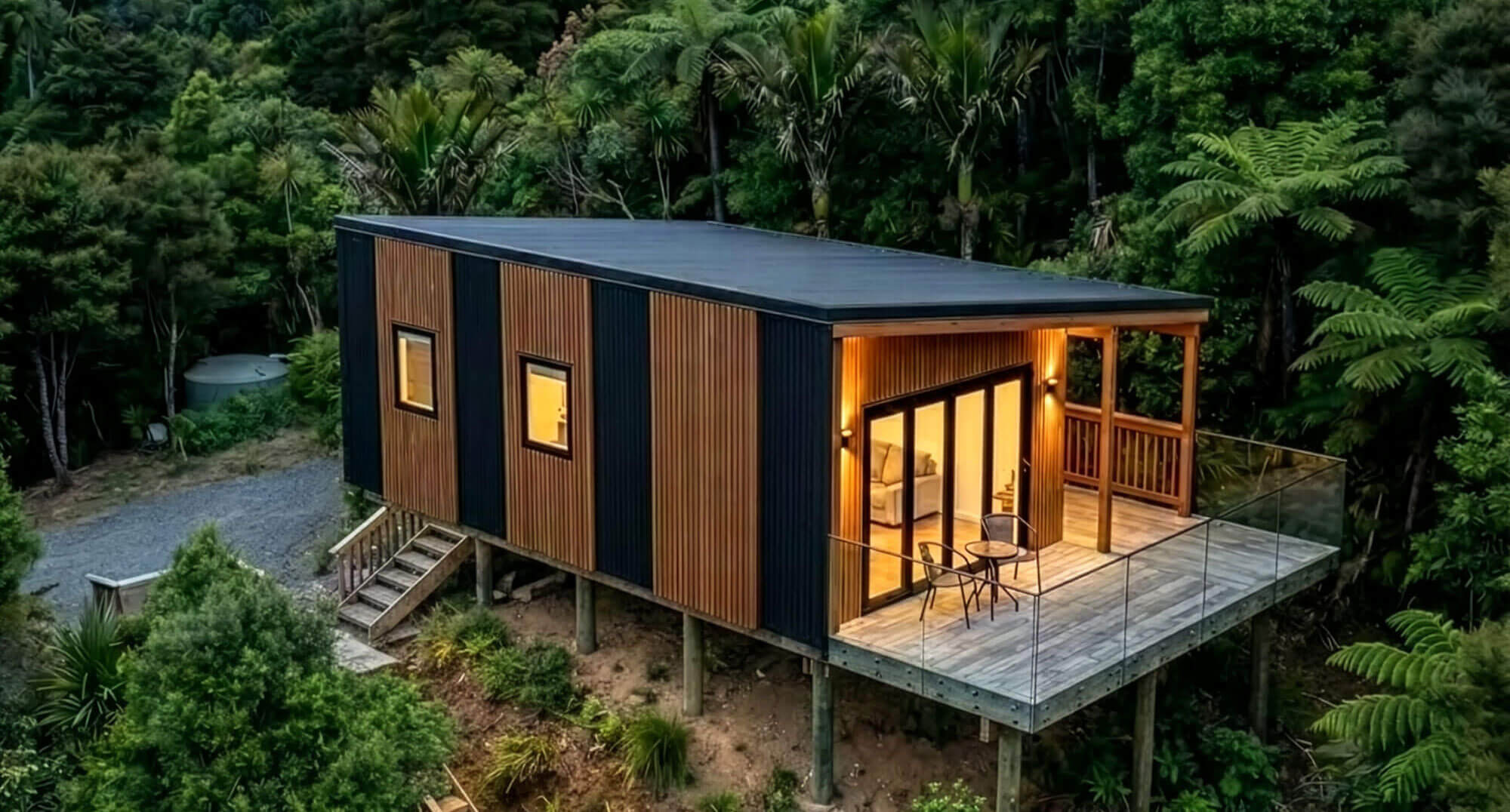 Modern cabin with large glass windows on a hillside surrounded by lush green trees and a winding wooden path.