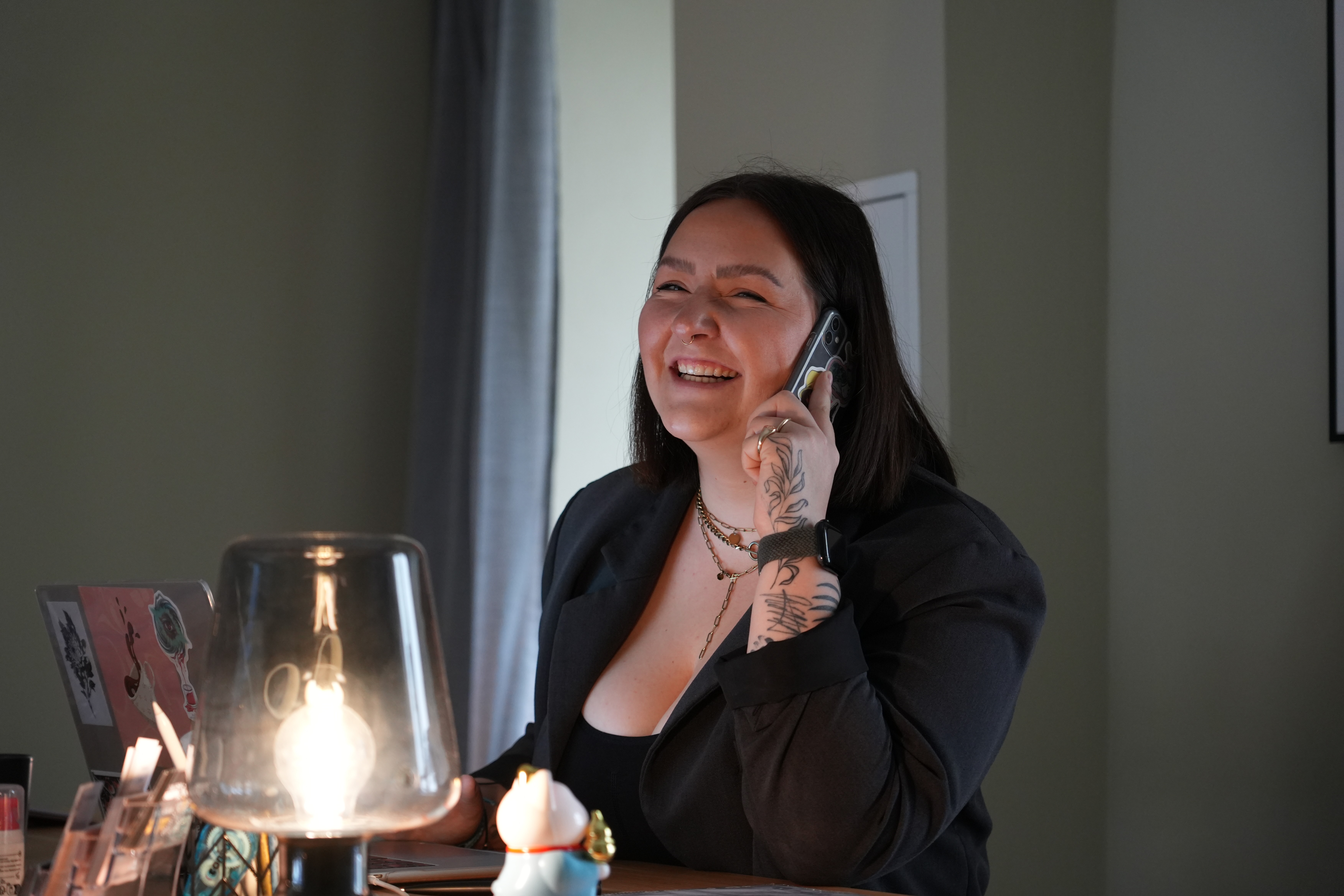 Eine Shop-Mitarbeiterin mit sichtbaren Tattoos am Unterarm sitzt an einem Tresen und telefoniert lächelnd. Auf dem Tisch stehen eine Lampe, ein Laptop mit Stickern und einige kleine Objekte. Die Szene vermittelt eine freundliche und serviceorientierte Atmosphäre in einem Tattoostudio.