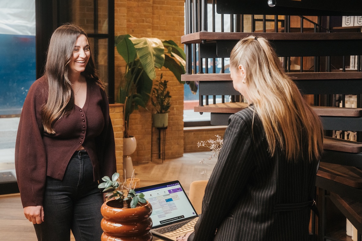 Two women talking in a salon reception area, with a laptop open on the desk