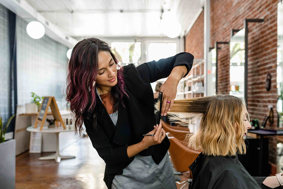 Hairstylist cutting a client's blonde hair in a bright salon with exposed brick walls