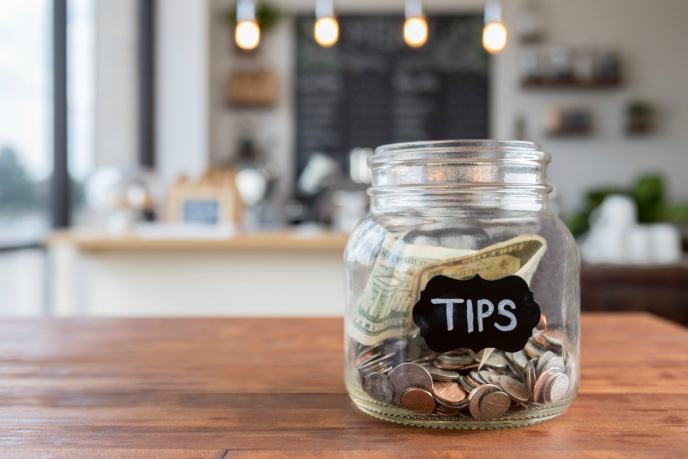 Glass tip jar full of coins and notes on a wooden counter