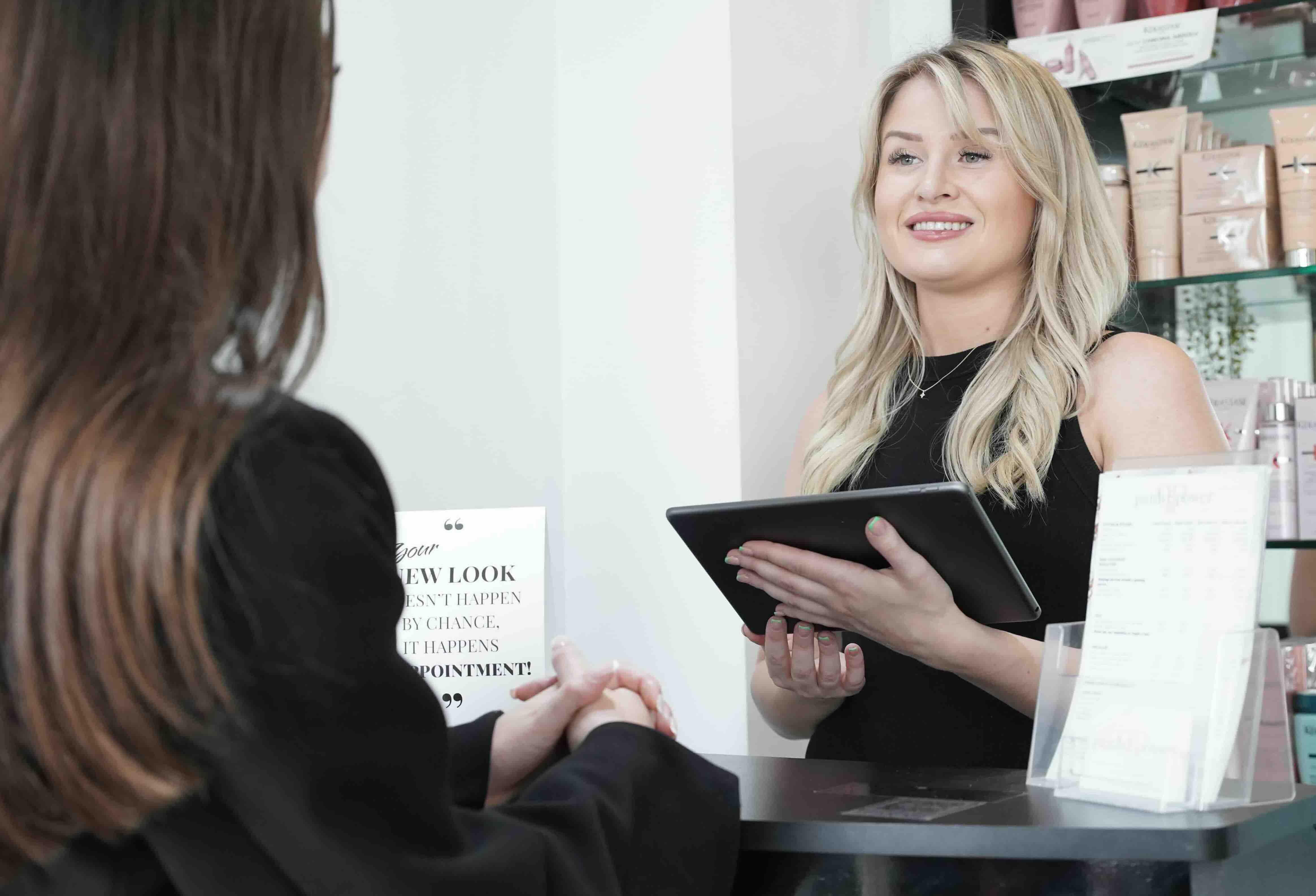 Salon receptionist holding a tablet and smiling at a client at the front desk