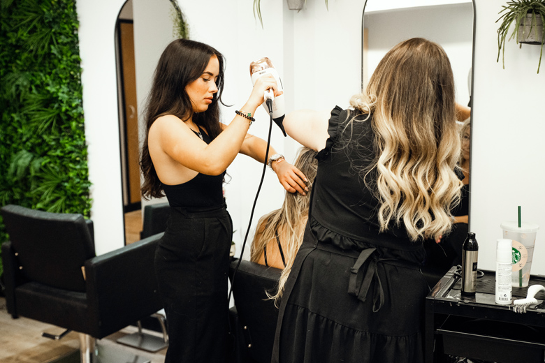 Two stylists blow-drying clients' hair in a salon with a plant wall