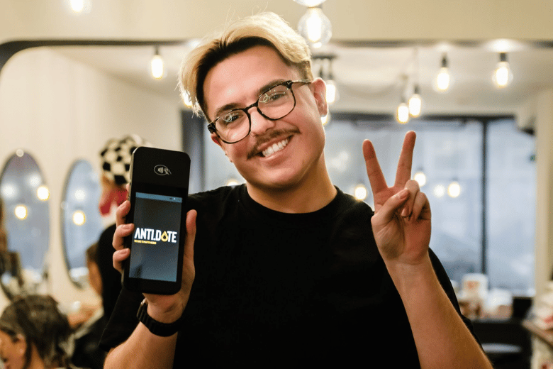 Smiling hairstylist holding up a phone and making a peace sign in a salon