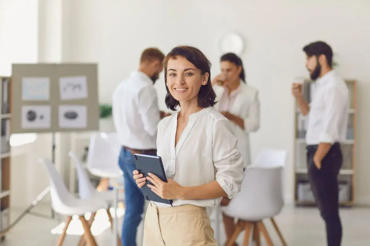 Woman holding a tablet while colleagues in background enjoy coffee.
