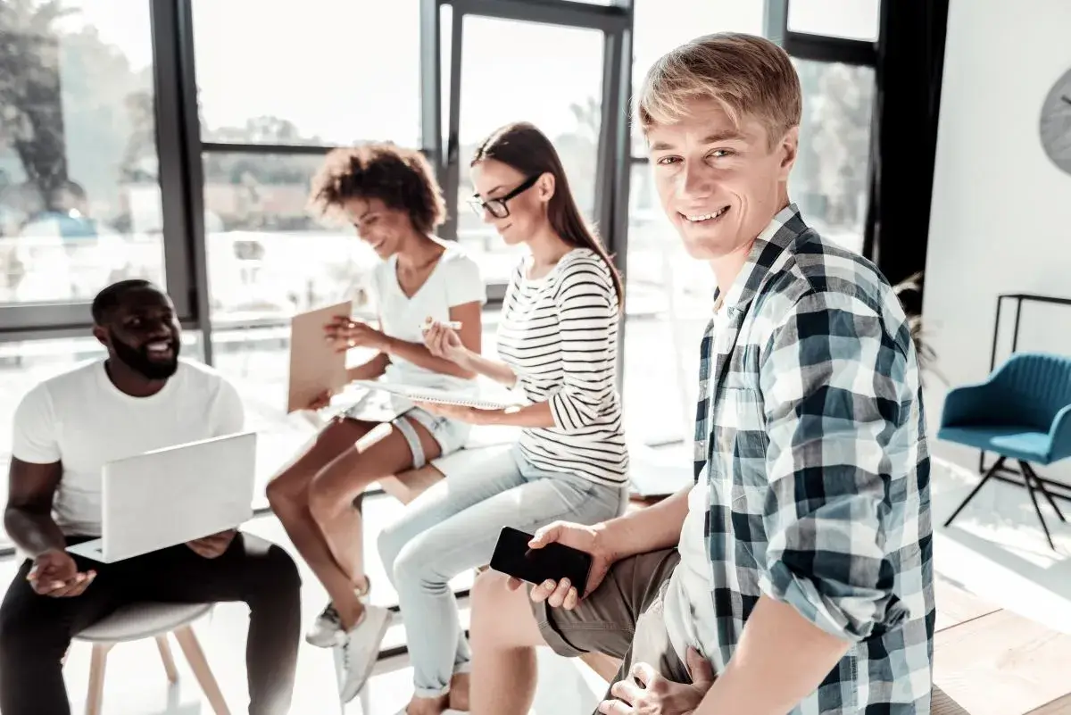 Four people conversing in a half-circle, while one male looks directly at the camera while holding his phone.