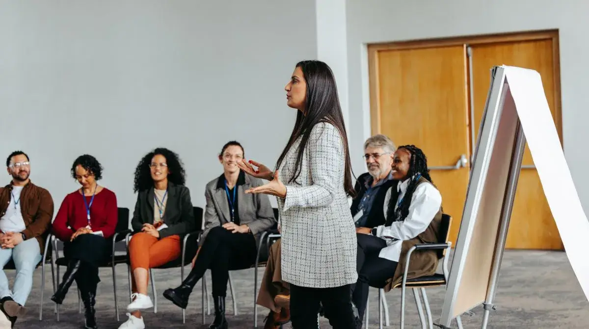 Group of people sitting and watching a woman speak in front of them.