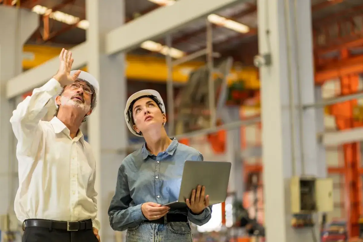 Man pointing at something and a woman holding a laptop looking up in a manufacturing facility.
