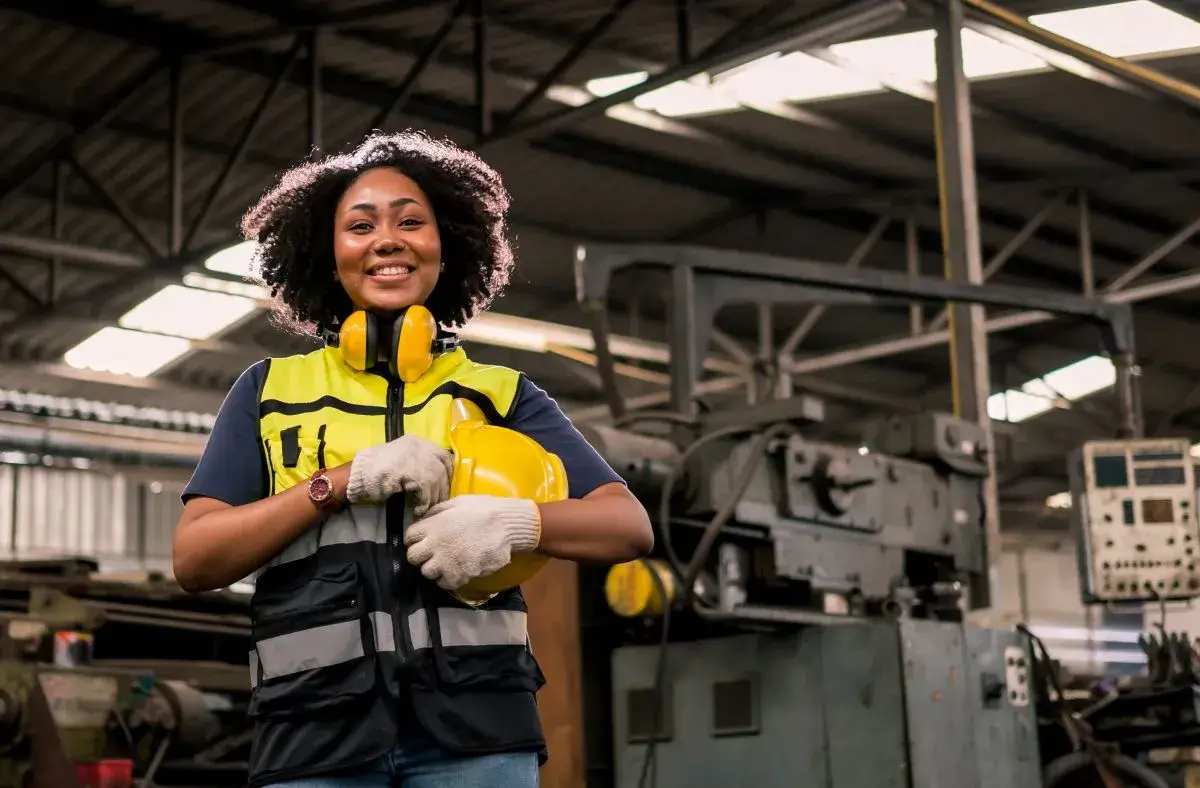 Woman in a manufacturing facility looking at the camera and holding her hard hat.