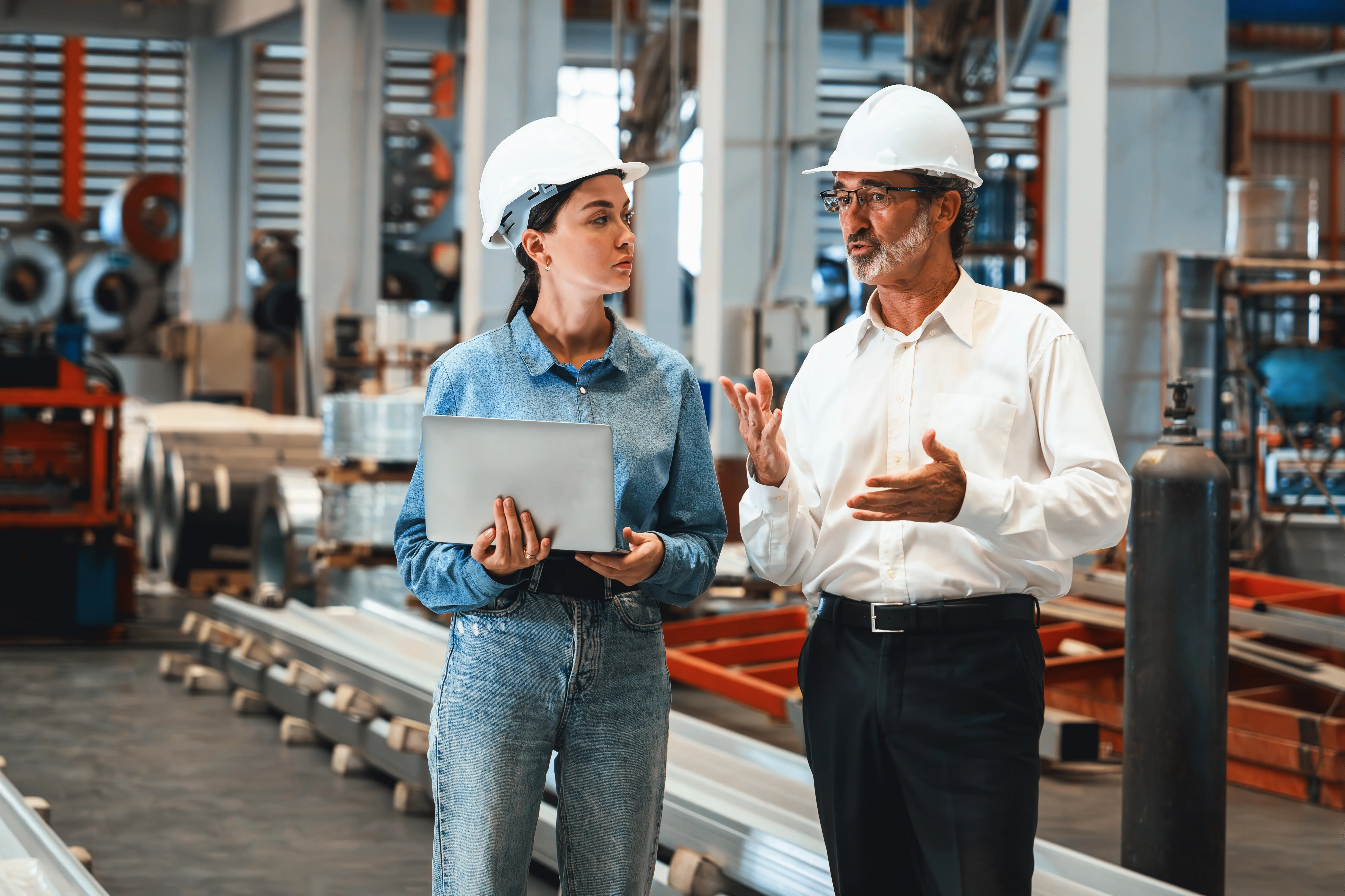 Man and woman standing in a manufacturing facility while both wearing white hard-hats and the woman is holding a silver laptop.
