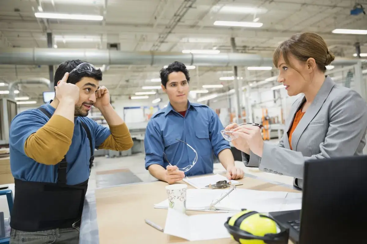 Two men and a woman in a gray blazer giving instructions on the safety equipment inside a manufacturing facility.