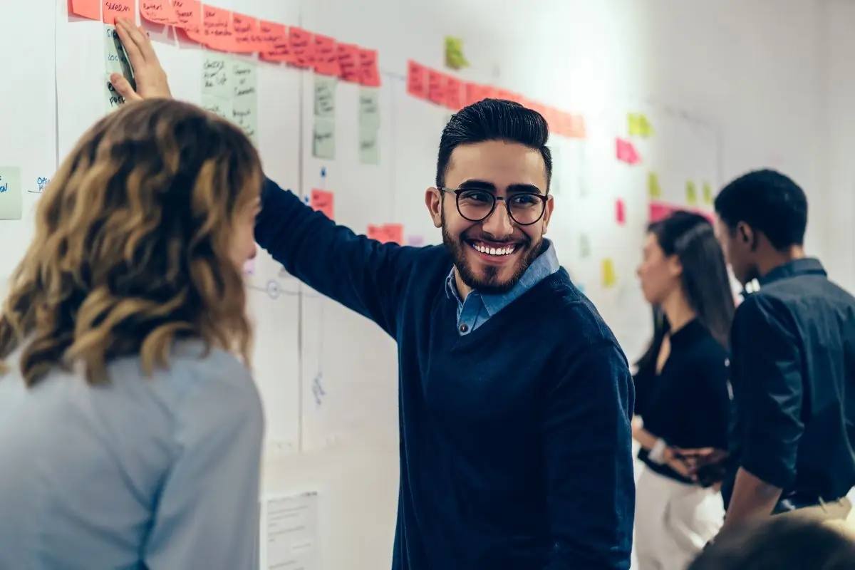 Man in collared shirt that's blue looking at someone while pointing to a sticky note on a whiteboard a smiling. A woman with brown hair and a gray vest is seen next to him also smiling, but only her side profile is shown.