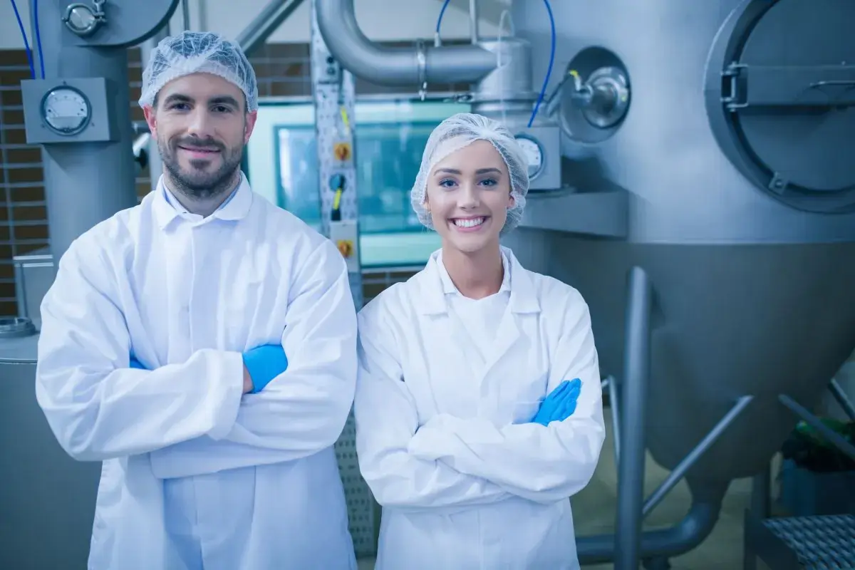 Man and woman wearing white lab coats and blue gloves inside a manufacturing facility while wearing hairnets and blue gloves.