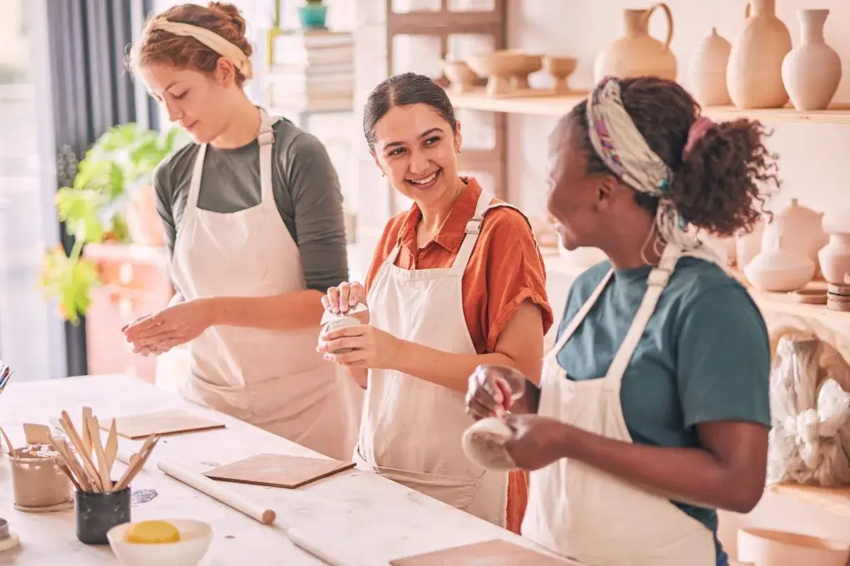 Three women in a studio crafting objects while smiling at each other.
