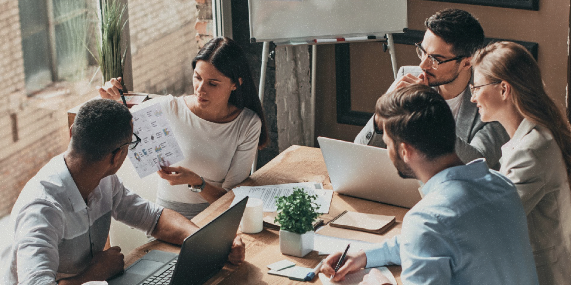 Five people sit around a table having a meeting in an office. A woman holds a document as they look to her.