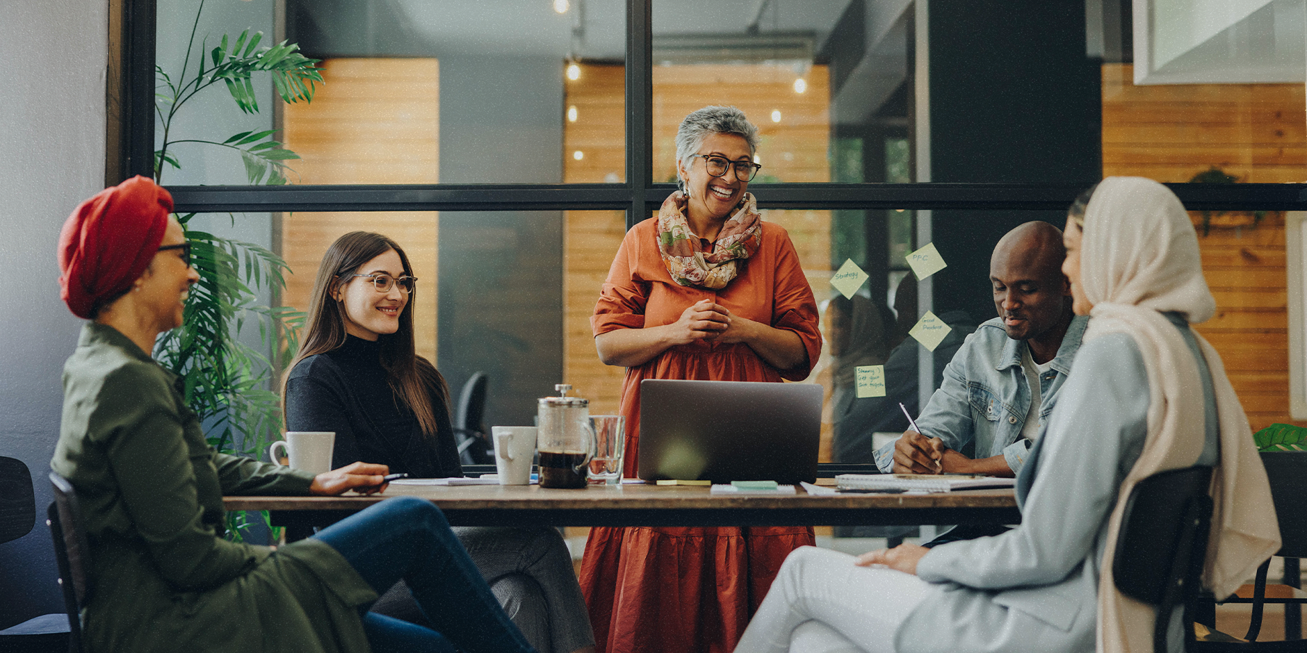 Five people sit around a table having a meeting in an office. A woman stands in the center and smiles as she looks at her coworkers.