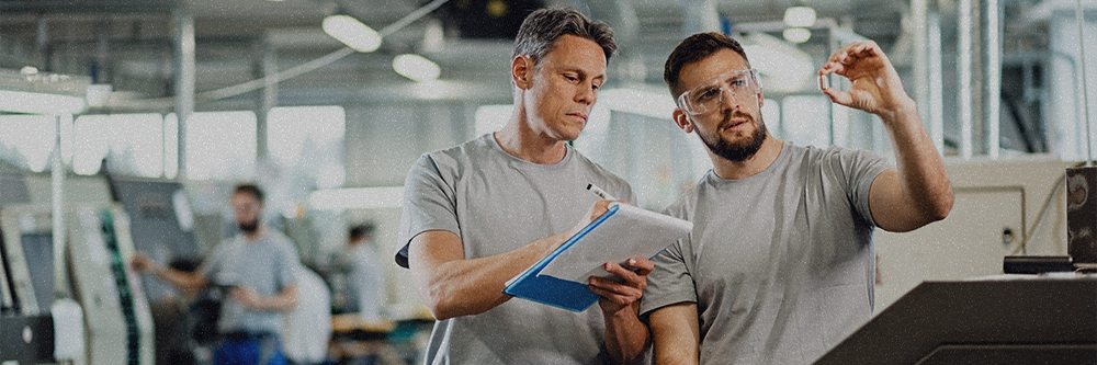 A man wearing safety goggles holds up a metal bit in a factory, while a second man writes on a clipboard.