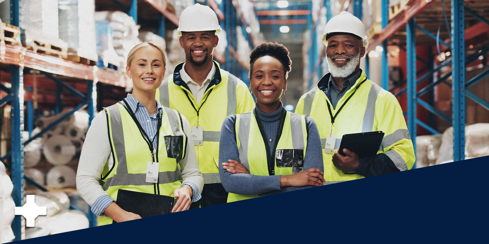 Four people in safety vests and hard hats stand in a warehouse and smile.