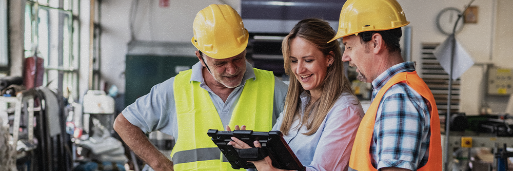 Two men in hard hats and safety vests stand on either side of a woman holding a tablet. All three look at the tablet in a work conversation.