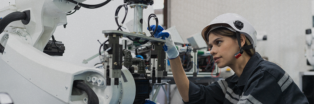 Woman in hard hat and safety gear uses gloved hands to examine a piece of machinery.